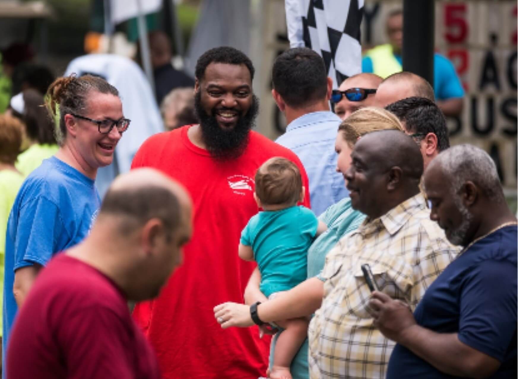 A joyful crowd gathering with a man holding a baby, sharing smiles and conversation.