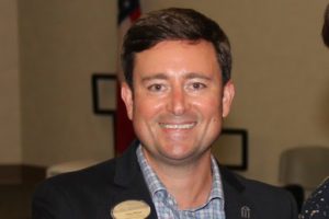 Smiling man wearing a suit jacket and checkered shirt indoors.