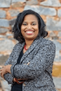 Smiling woman in a gray blazer stands confidently outdoors.