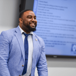 A confident man in a blue suit smiling during a presentation.