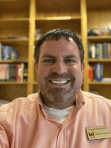 Smiling man in peach shirt stands in front of bookshelf.