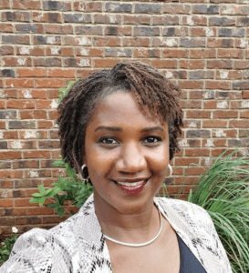 Smiling woman with short curly hair in front of a brick wall.