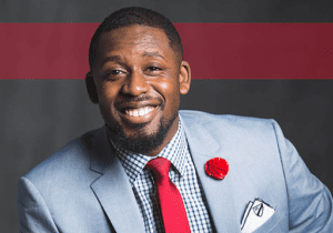 Smiling man in a light blue suit with a red tie and floral lapel pin.