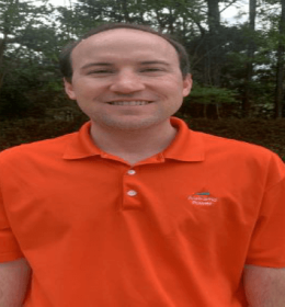 Man in orange polo shirt standing outdoors with trees in the background.