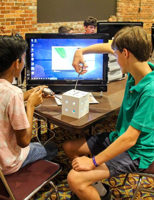 Two boys collaborating on a 3D design project using a computer and a small model.